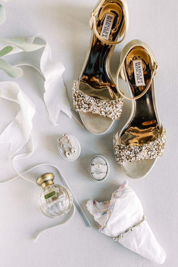 Wedding flat lay detail photo on a white textured backdrop with bejeweled Badgley Mischka sandals, white ribbon, and Tocca perfume bottle