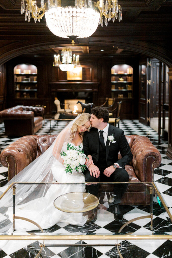 Bride and groom sit on a tufted brown leather couch inside a dark paneled room with a black and white checkered floor