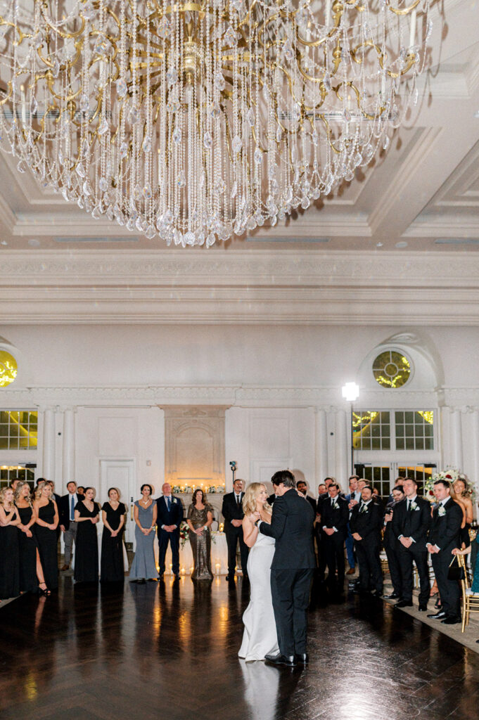 Surrounded by onlookers, bride and groom dance their first dance under a crystal chandelier in the ballroom at the Park Chateau