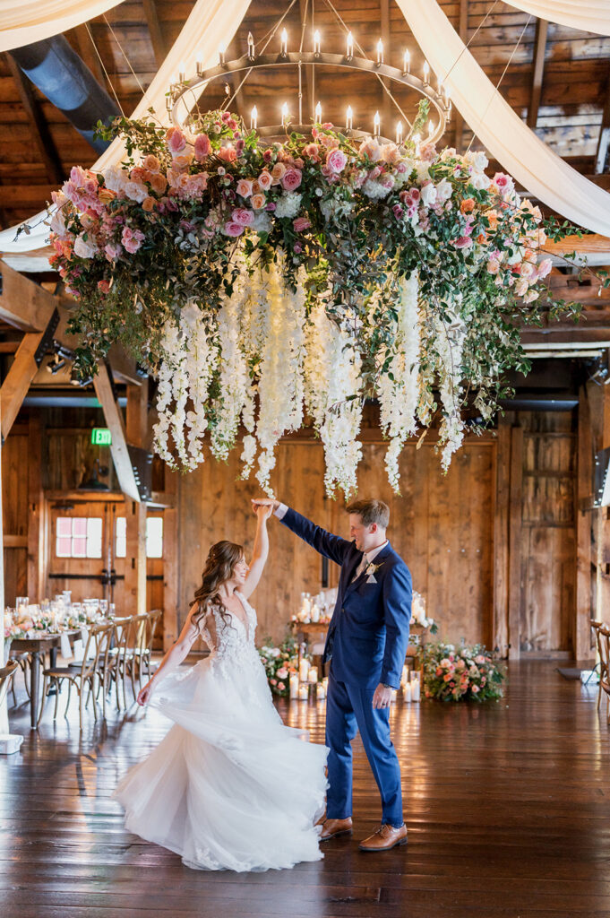 Huge floral installation with white and pink flowers hangs from the ceiling of a wedding reception space over a bride and groom dancing