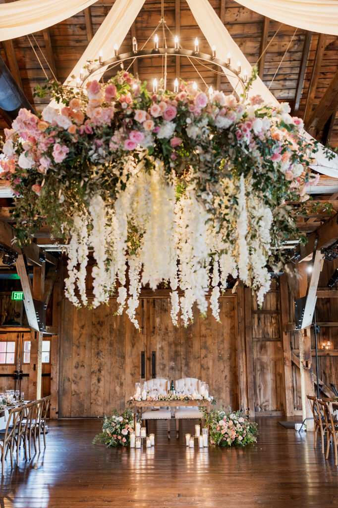 Huge floral installation with white and pink flowers hangs from the ceiling of a wedding reception space over the sweetheart table
