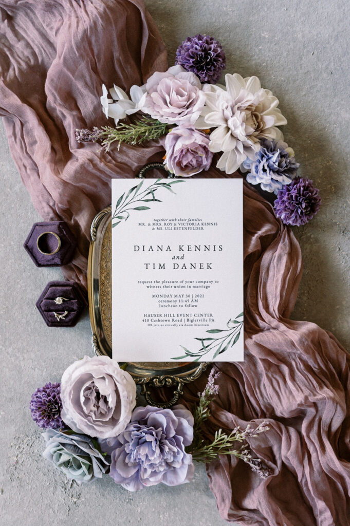 Wedding detail flat lay of simplistic wedding invitation on a silver tray surrounded by purple florals and purple linen fabric