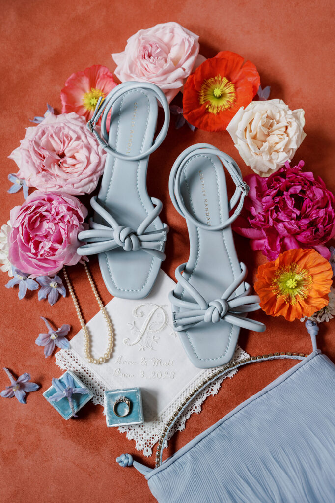Brightly colored wedding detail flat lay with pastel blue strappy bridal sandals surrounded by orange and pink florals and a periwinkle clutch