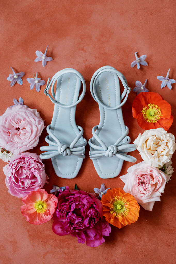 Brightly colored wedding detail flat lay with pastel blue strappy bridal sandals surrounded by orange and pink florals