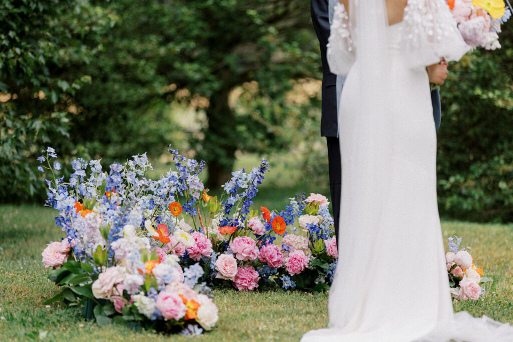 Bride and groom stand in front of an orange pink white and blue floral installation that sits on the ground as a backdrop for their simple wedding ceremony