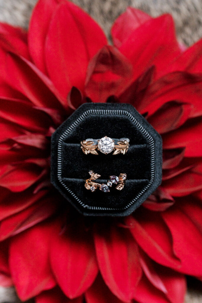 Black velvet ring box sits on a vibrant red flower and holds two intricately designed gold and diamond rings 