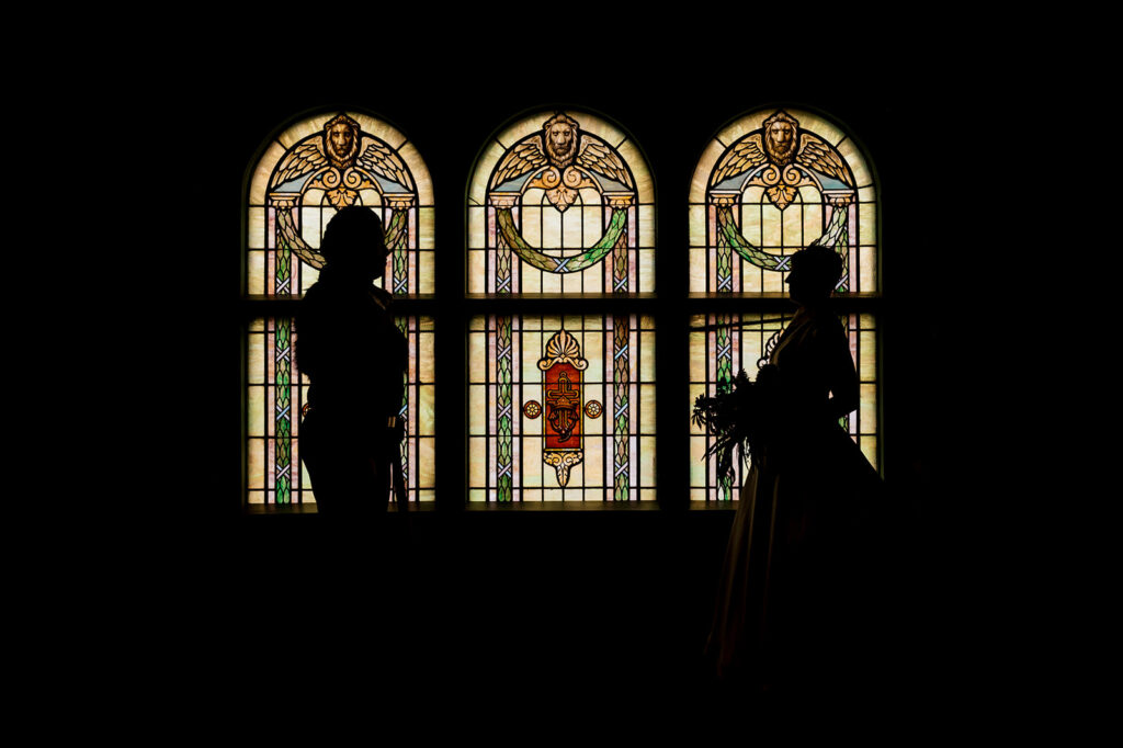 Bride and groom face each other silhouetted against a wall of three arched stained glass windows