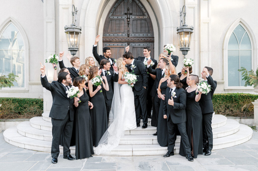 Bridal party wearing all black surrounds bride and groom on the steps in front of The Chapel at The Park Chateau wedding venue in East Brunswick NJ