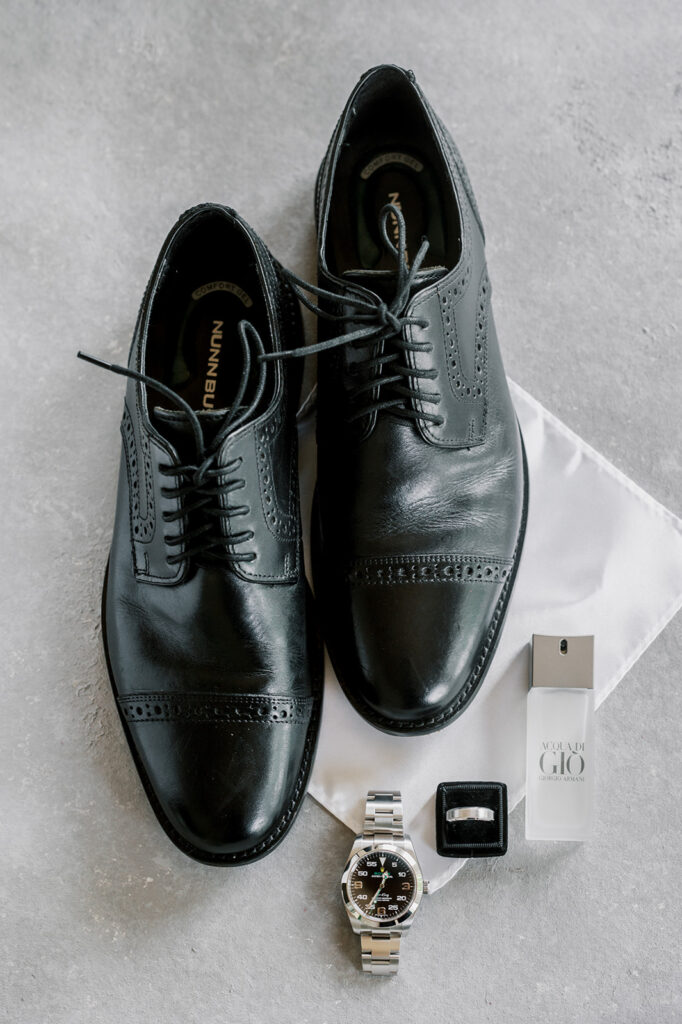 Simple black and white wedding detail flat lay of a groomsman's black leather shoes, Acqua Di Gio By Giorgio Armani cologne, a silver watch and a silver wedding band in a black velvet ring box
