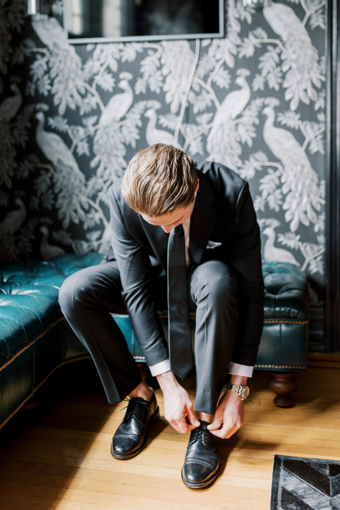 Groom sits on a jewel toned blue leather bench and leans over to tie his black leather shoes