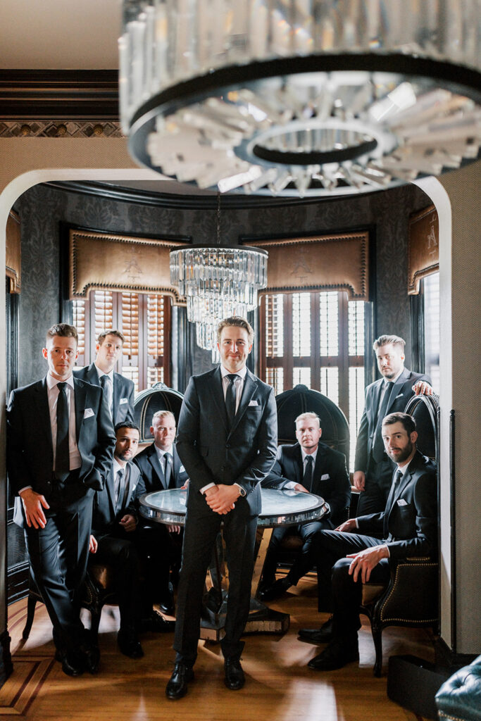 Groom poses surrounded by seven groomsmen inside a tasteful and moody grooms suite at the Willows at Ashcombe wedding venue