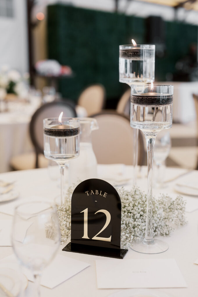 Simplistic black and white table setting for wedding with three glass floating candle holders and black acrylic table number surrounded by baby's breath flowers