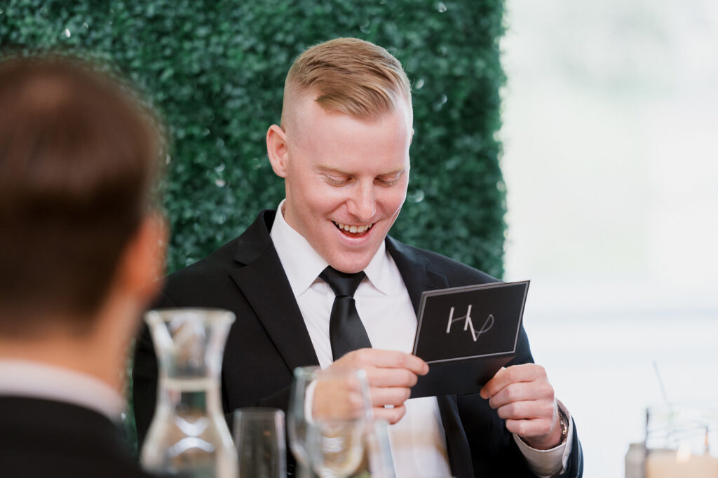 Groomsman sits in a chair and grins as he read a personalized thank you note from the bride and groom