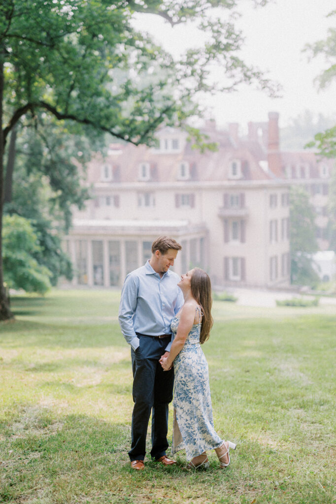 Engaged couple in coordinating blue engagement photo outfits hold hands and look at each other with Winterthur museum in the background for their engagement photos in Wilmington, DE