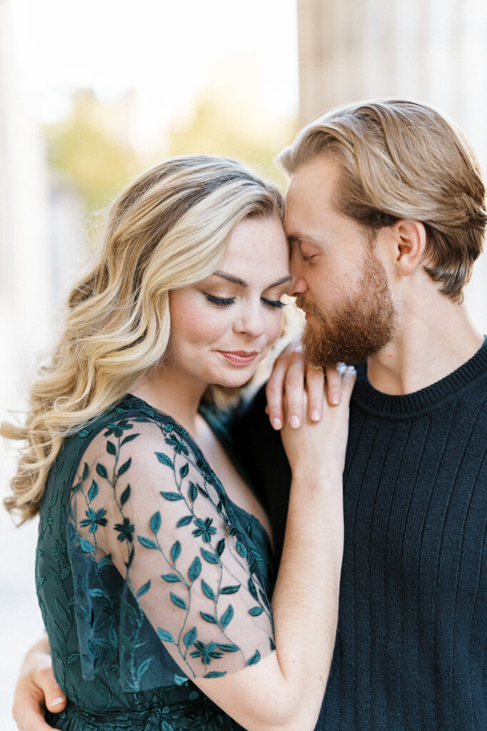 Blonde woman in emerald green dress looks down as her fiancé rests his forehead on hers
