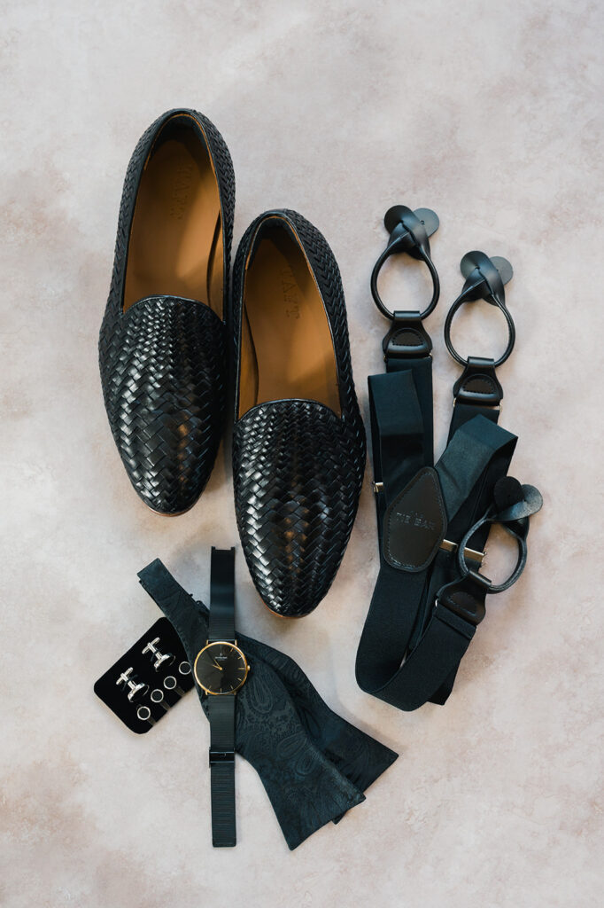 Woven black loafers sit next to black suspenders, black watch, black bowtie, and set of silver cuff links