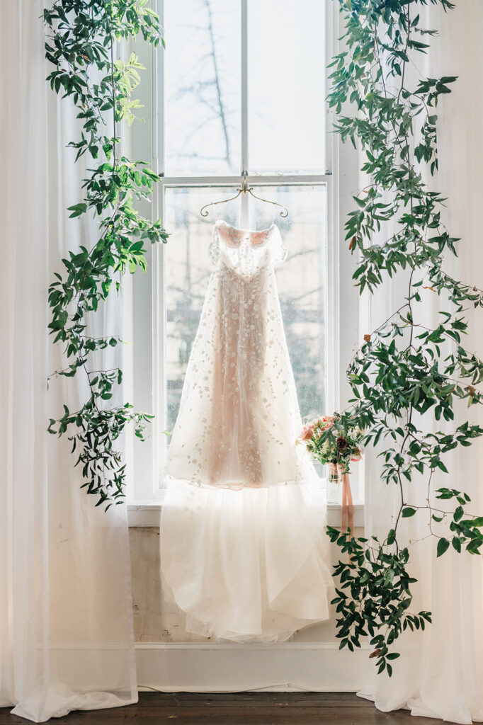 Wedding dress hangs on a delicate hanger in front of a tall window framed by hanging greenery and white curtains