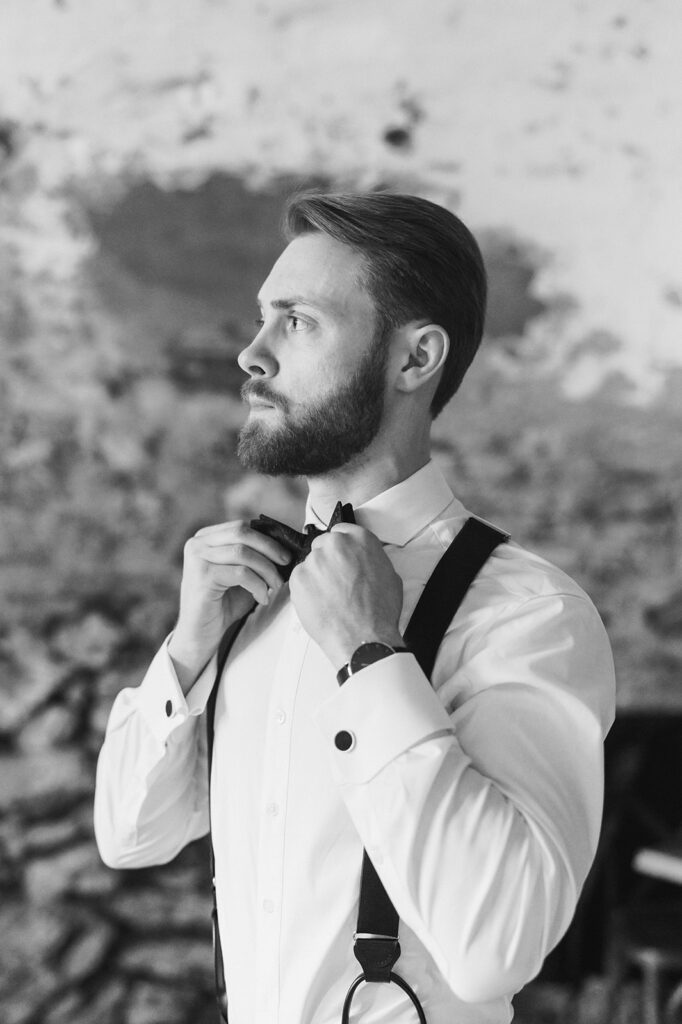 Groom wearing a white dress shirt and suspenders looks away as he fixes his black bowtie 