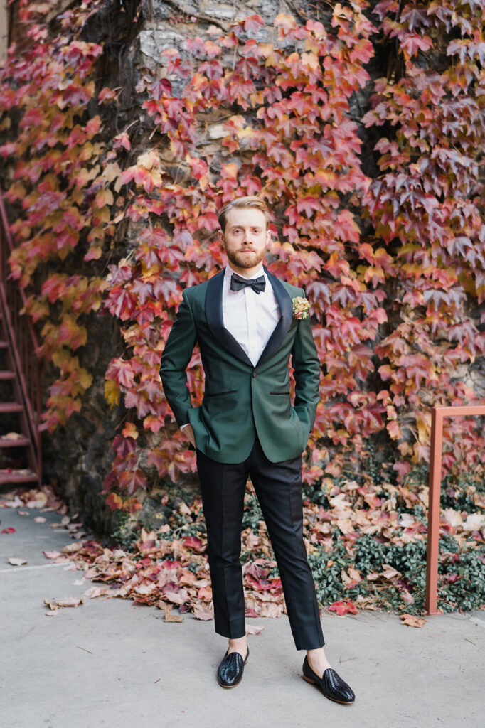 Groom poses with hands in his pockets wearing an emerald green dinner jacket with a black shawl collar and black loafers