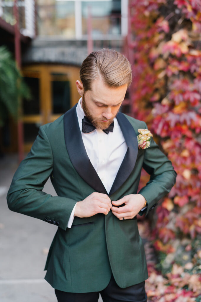 Groom looks down as he buttons his emerald green dinner jacket with a black shawl collar
