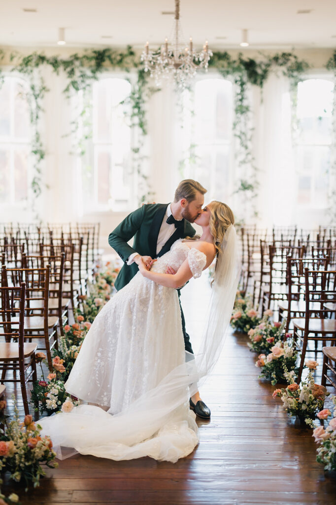Groom in emerald green dinner jacket dips his bride who is wearing a white dress with floral appliqués and cap sleeves in the aisle of their wedding ceremony space at Excelsior