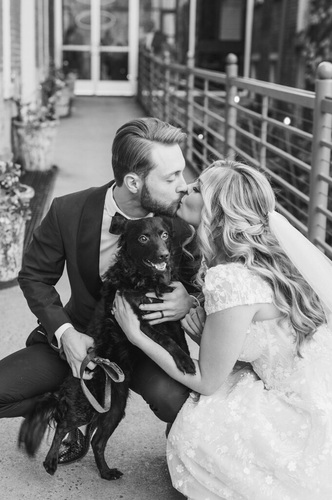 Bride and groom kiss as they pose with their black Mudi dog outside of Excelsior wedding venue in downtown Lancaster PA