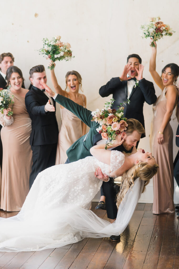 Groom dips his bride in front of cheering bridal party as she throws her head back and laughs