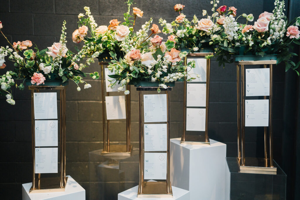 Four metal podiums topped with floral arrangements hold hanging cards with seating assignments for wedding guests