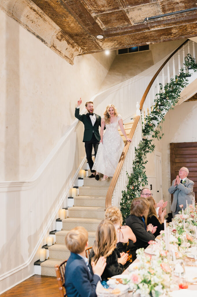 Bride and groom descend tall curved staircase into the Excelsior venue reception space where their guests are seated