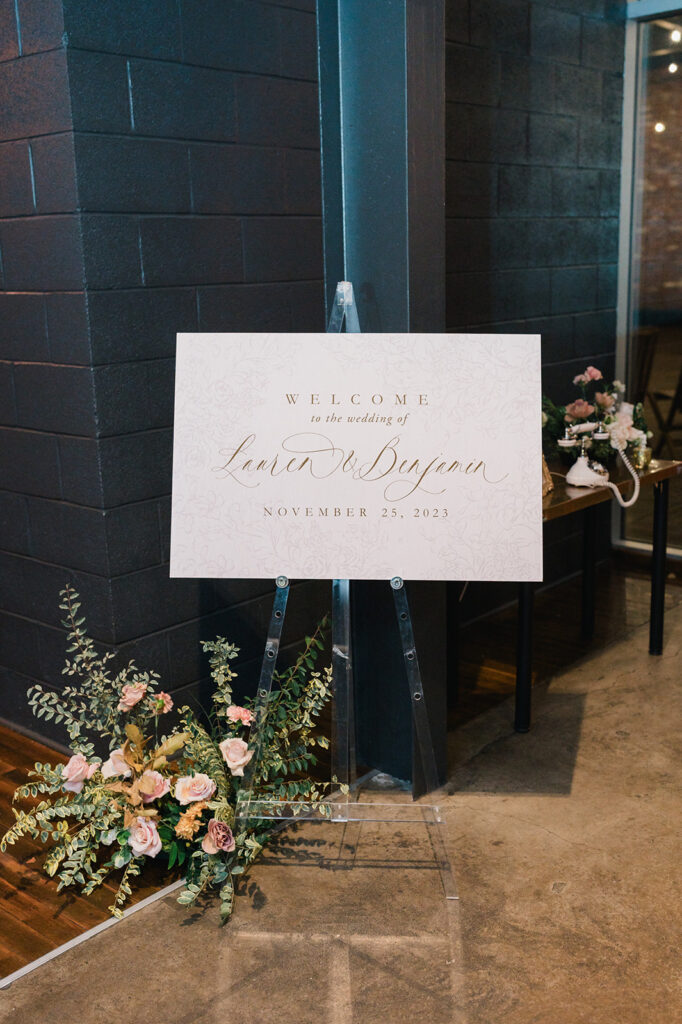 Wedding welcome sign sits on a clear acrylic easel next to a green and pink floral arrangement