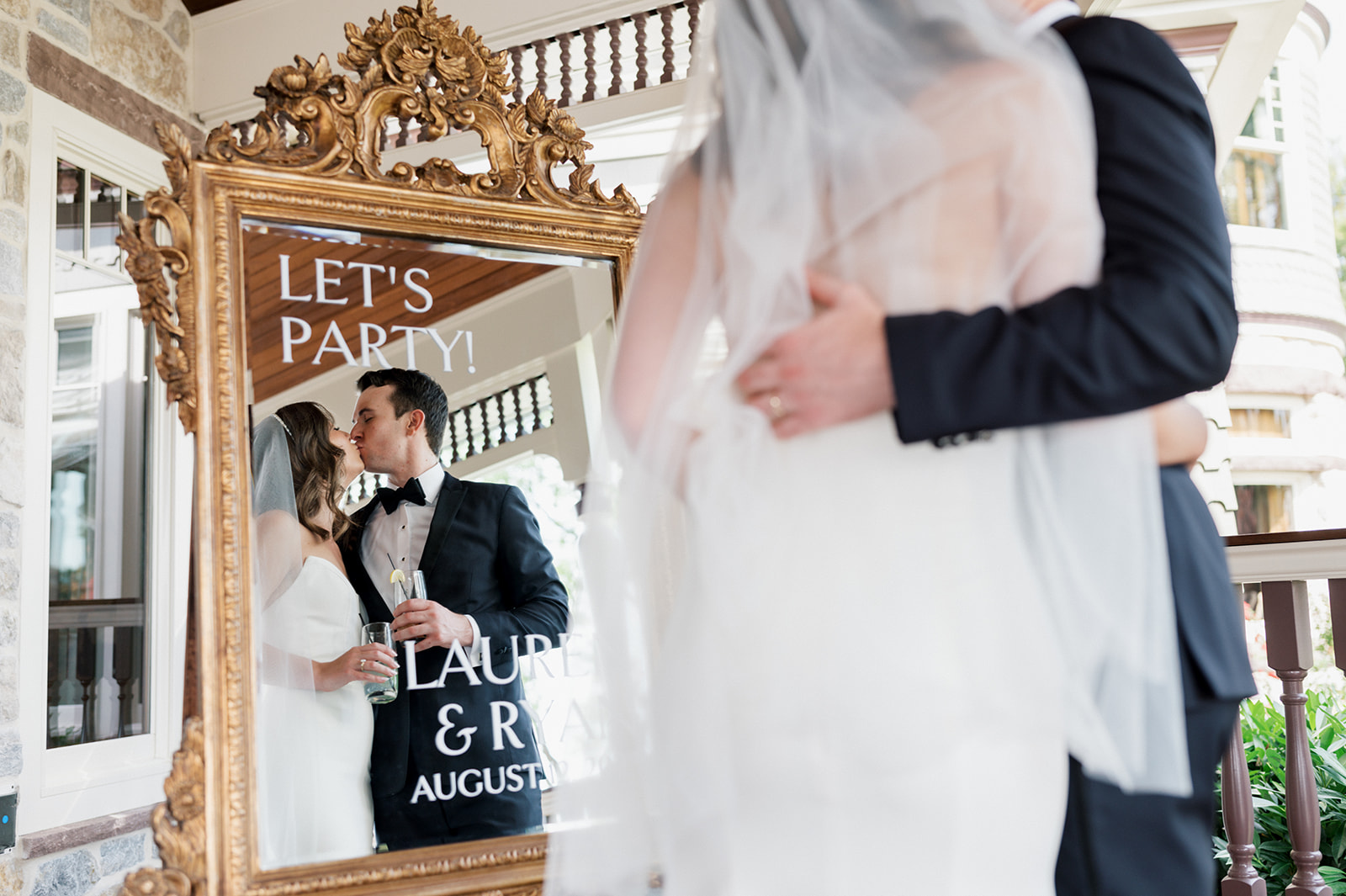 Bride and groom kiss in front of a gold framed mirror that reads "Let's Party"