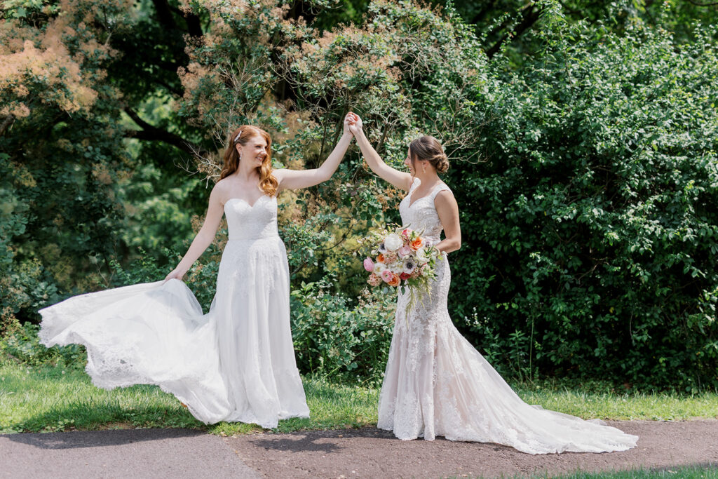 two brides in white dresses hold hands as they twirl in front of a backdrop of dense greenery at the John James Audubon Center
