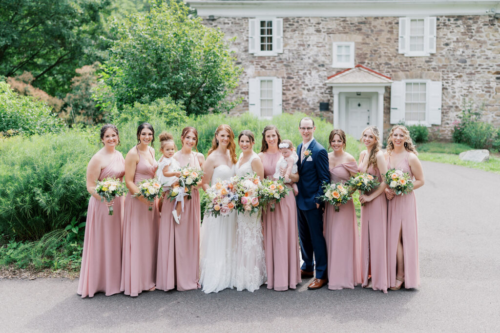 Seven Bridesmaids wearing dusty rose dresses and one bridesman wearing a navy suit surround two brides on their wedding day