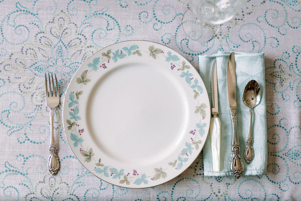 Simple blue and green floral rimmed white plate sits on blue and white tablecloth flanked by silverware