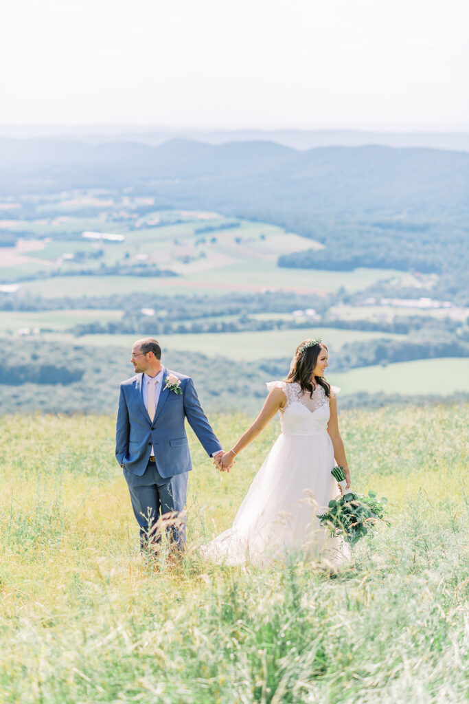 Bride and groom hold hands and look opposite directions as they stand on a hillside overlooking a grassy valley below