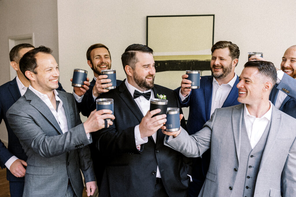 Six groomsmen in gray suits hold up navy blue Yeti thermoses