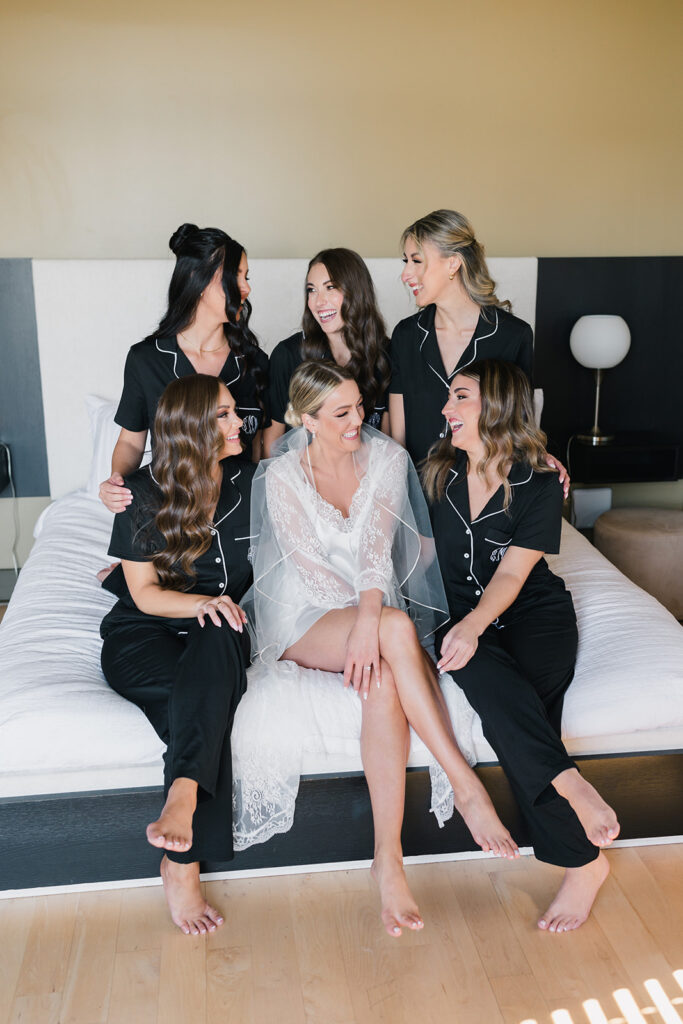 Bride in white lace robe sits on bed surrounded by 5 bridesmaids in black pajama sets