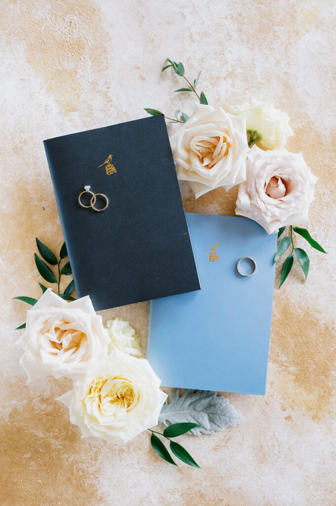 Wedding flat lay detail photo on a cream textured backdrop with two blue vow books and white rose cuttings