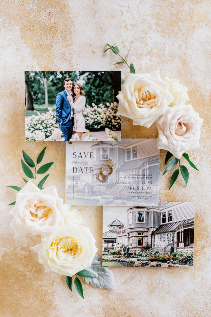 Wedding flat lay detail photo on a cream textured backdrop with three save the date stationary pieces, wedding rings, and white rose cuttings