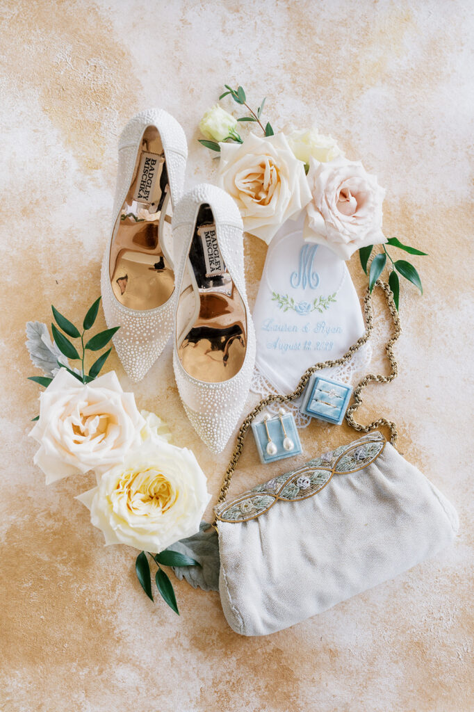 Wedding flat lay detail photo on a cream textured backdrop with white pearl Badgley Mischka shoes, vintage clutch purse, and white rose cuttings