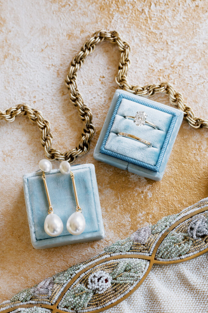 Wedding flat lay detail photo on a cream textured backdrop with tiffany blue velvet ring box holding wedding rings and white and gold pearl earrings