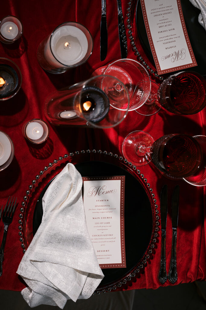 Close up image of red and white wedding dinner menu and white napkin sitting on a clear beaded edge charger surrounded by black, red, and white decor