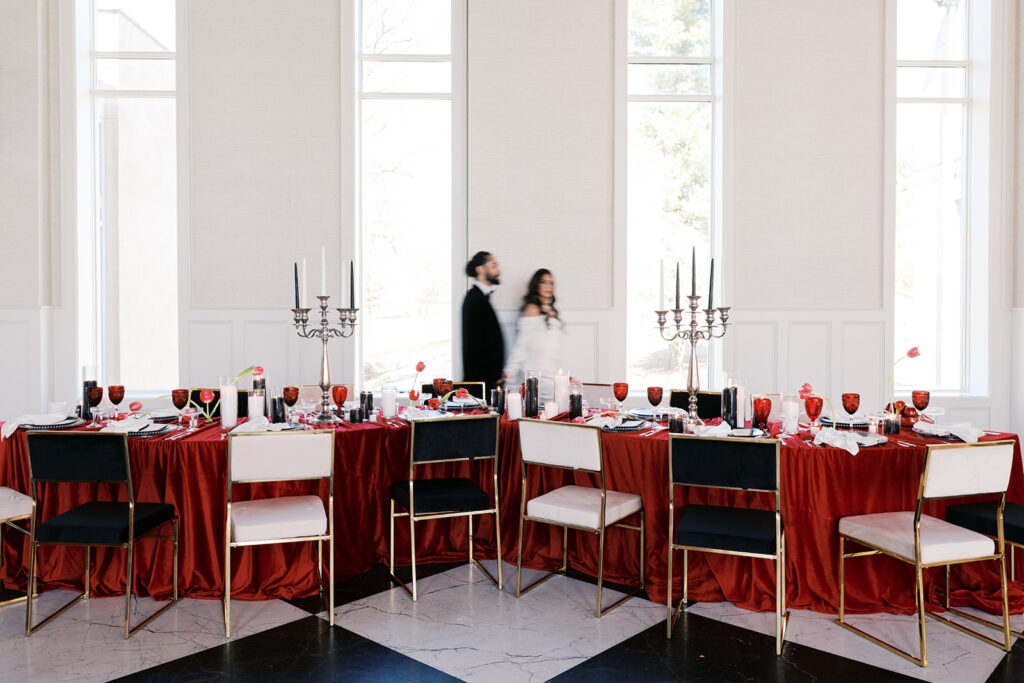 Bride and groom walk behind S-shaped banquet table covered in red linen surrounded by alternating black and white chairs on a checkerboard floor at Elkins Estate in PA