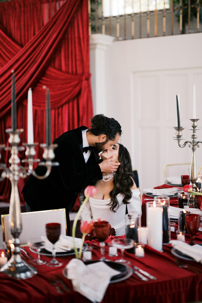 Groom kisses bride as she sits beside banquet table covered in red linen and black and white wedding reception decor at Elkins Estate | Lindsey Ford Photography