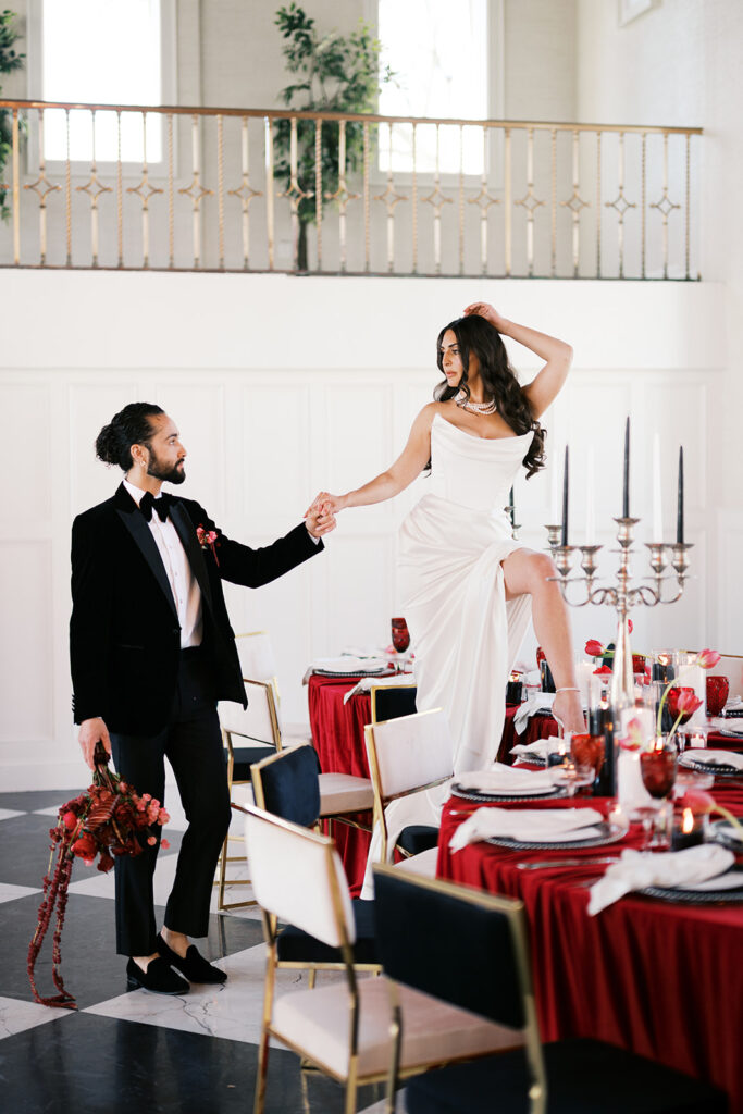 Groom helps bride step up onto curved banquet table covered in red linen and black and white wedding reception decor at Elkins Estate | Lindsey Ford Photography