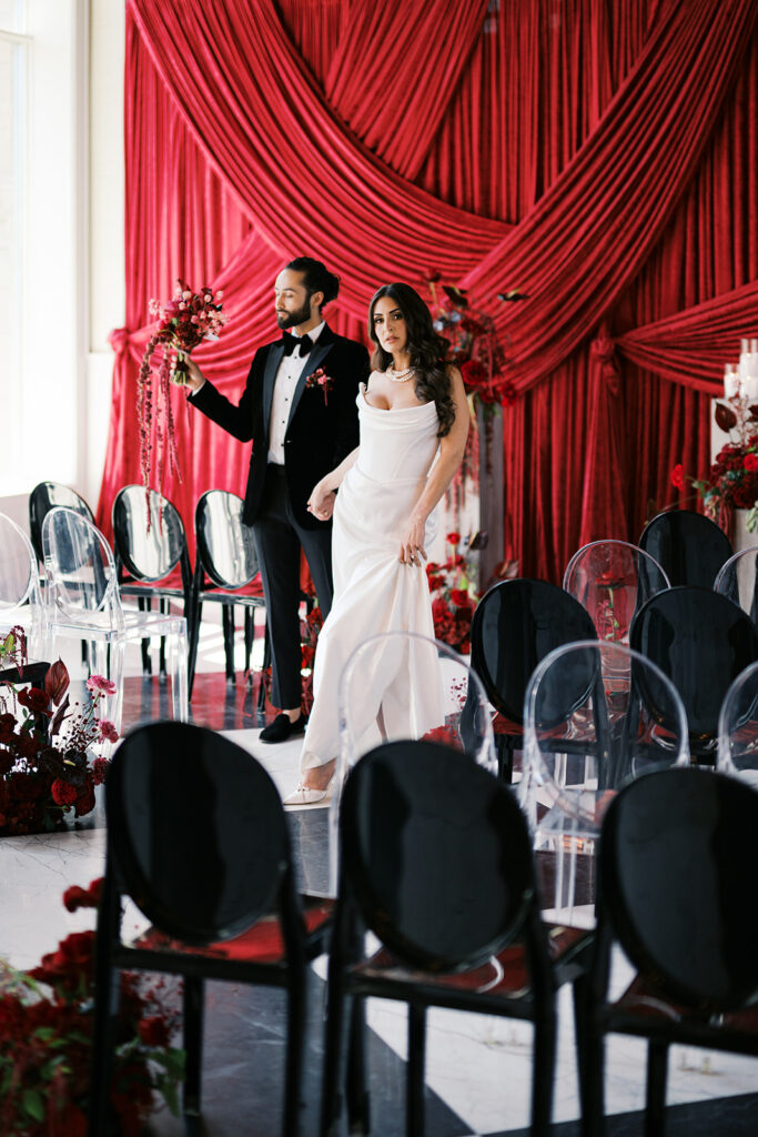 Bride and groom pose together against wedding ceremony backdrop of dark red draping surrounded by all red wedding flowers at Elkins Estate in PA