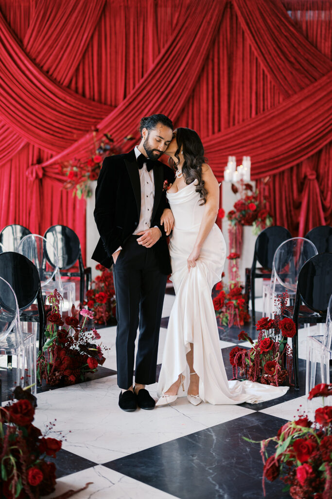 Bride and groom pose together against wedding ceremony backdrop of dark red draping surrounded by all red wedding flowers at Elkins Estate in PA