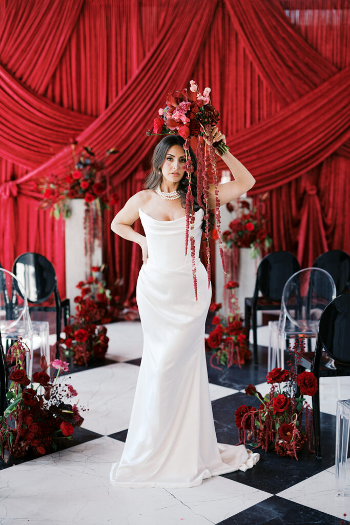 Bride poses holding an all red bouquet over her head against wedding ceremony backdrop of dark red draping at Elkins Estate in PA