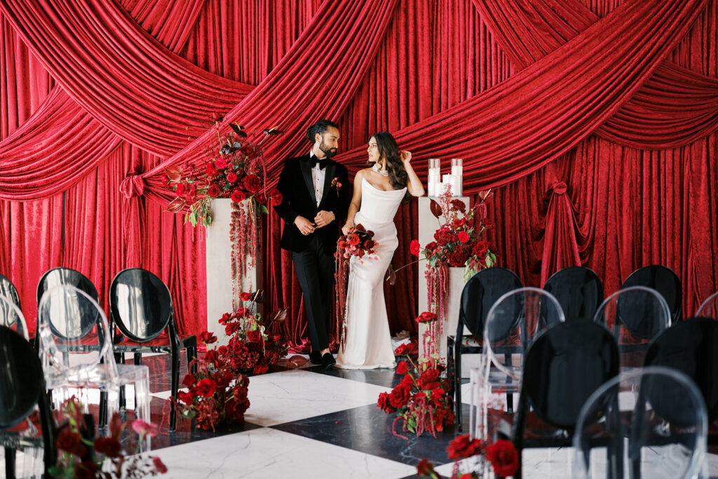 Bride and groom pose together against wedding ceremony backdrop of dark red draping surrounded by all red wedding flowers at Elkins Estate in PA