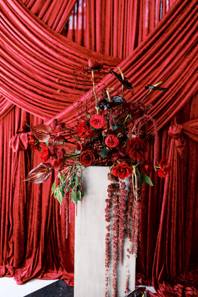 Large red wedding flower arrangement sits on white platform in front of ceremony backdrop of dark red draping at Elkins Estate in PA